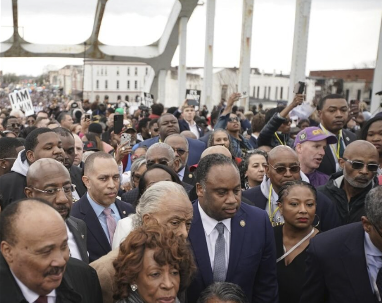A large crowd of predominantly Black men and women march shoulder-to-shoulder on a cloudy day at the Edmund Pettis Bridge in Selma, Alabama. The white steel supports of the bridge are shown overhead, and among the faces in the crowd is U.S. Representatives Hakeem Jeffries, who is wearing a dark suit with a light blue shirt and pink tie. Also present are U.S. Representative Maxine Waters, in a tweed coat, as well as civil rights leaders Al Sharpton, partially obscured behind Waters, and Jesse Jackson, who is seated in a wheelchair with sunglasses on and his head down. At right is ACLU President Deborah Archer, who is seen in a dark top with a light purse strap across her chest.