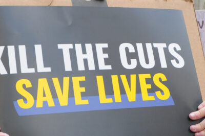 A demonstrator holds up a sign during a protest that reads "KILL THE CUTS, SAVE LIVES".