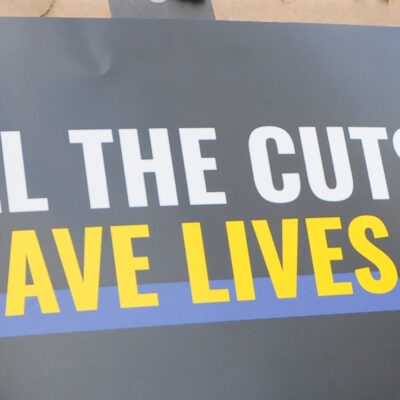 A demonstrator holds up a sign during a protest that reads "KILL THE CUTS, SAVE LIVES".