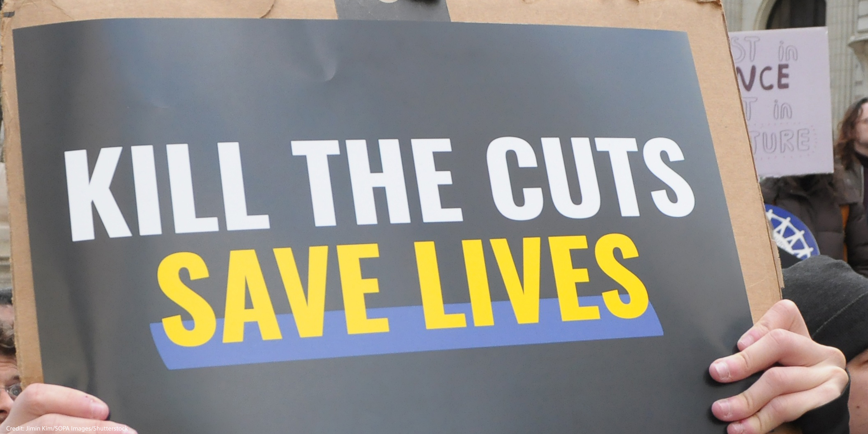 A demonstrator holds up a sign during a protest that reads "KILL THE CUTS, SAVE LIVES".