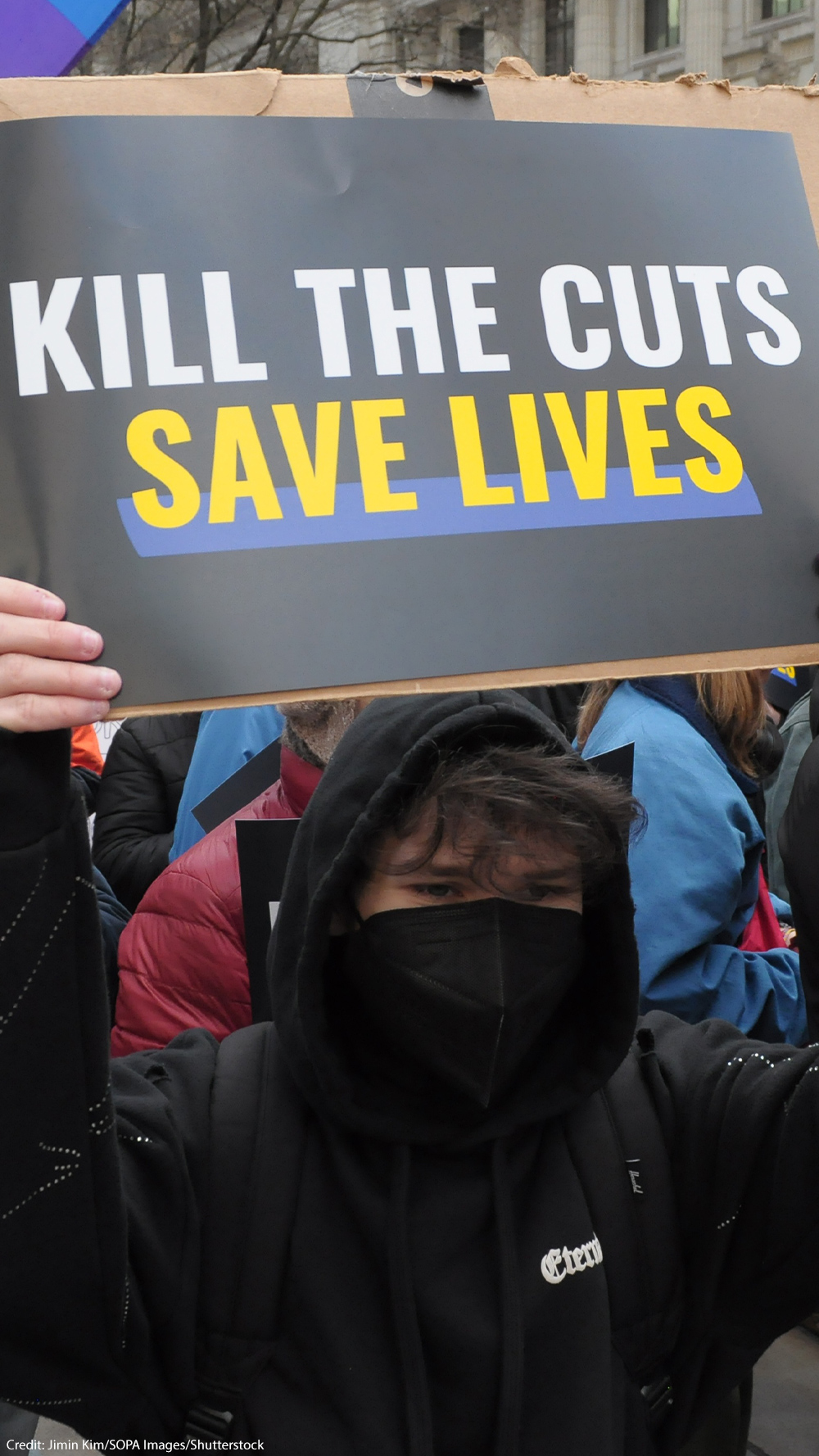 A masked demonstrator in a black hoodie holds up a sign during a protest that reads "KILL THE CUTS, SAVE LIVES".
