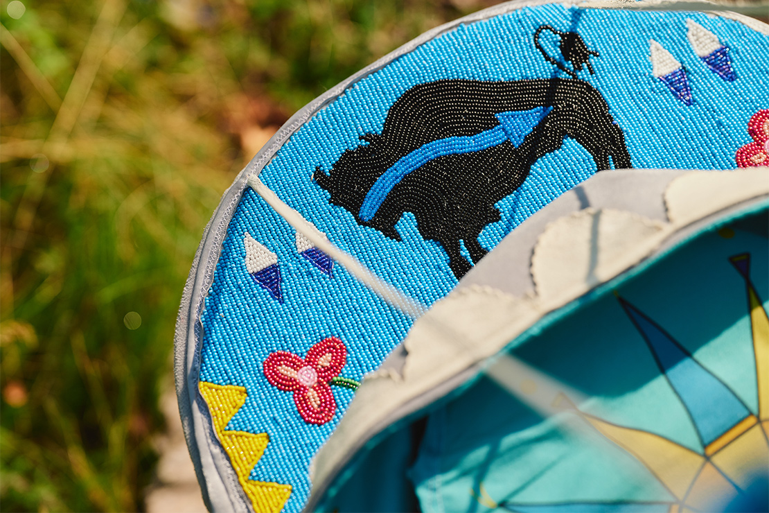 Close-up of intricate turquoise beadwork on an Indigenous cradleboard, featuring a black bison with blue detailing, geometric arrow-like patterns, and a red floral design, with part of the cradleboard’s painted blue interior visible in the foreground.