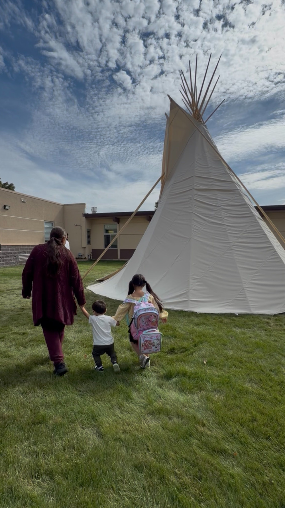An adult walks hand-in-hand with two children across a grassy lawn toward a large white teepee beside a building, under a wide, cloud-filled sky.