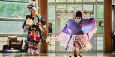 Two Indigenous dancers in colorful traditional regalia perform indoors; one wears a flowing purple shawl with long pink-and-white fringe, while the other wears a feathered headdress and holds a feather fan, with sunlight streaming through large windows behind them.