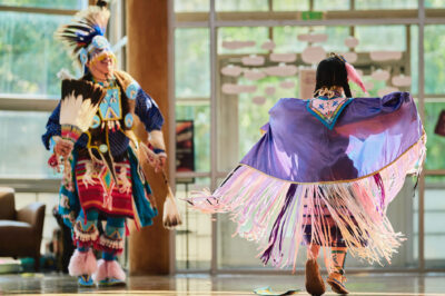 Two Indigenous dancers in colorful traditional regalia perform indoors; one wears a flowing purple shawl with long pink-and-white fringe, while the other wears a feathered headdress and holds a feather fan, with sunlight streaming through large windows behind them.