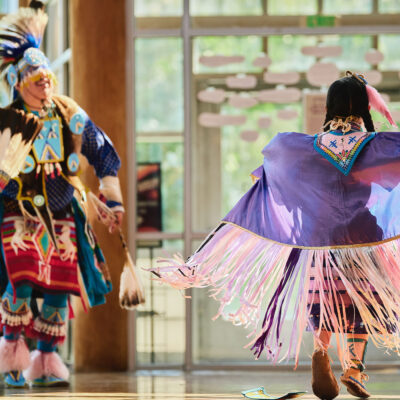 Two Indigenous dancers in colorful traditional regalia perform indoors; one wears a flowing purple shawl with long pink-and-white fringe, while the other wears a feathered headdress and holds a feather fan, with sunlight streaming through large windows behind them.