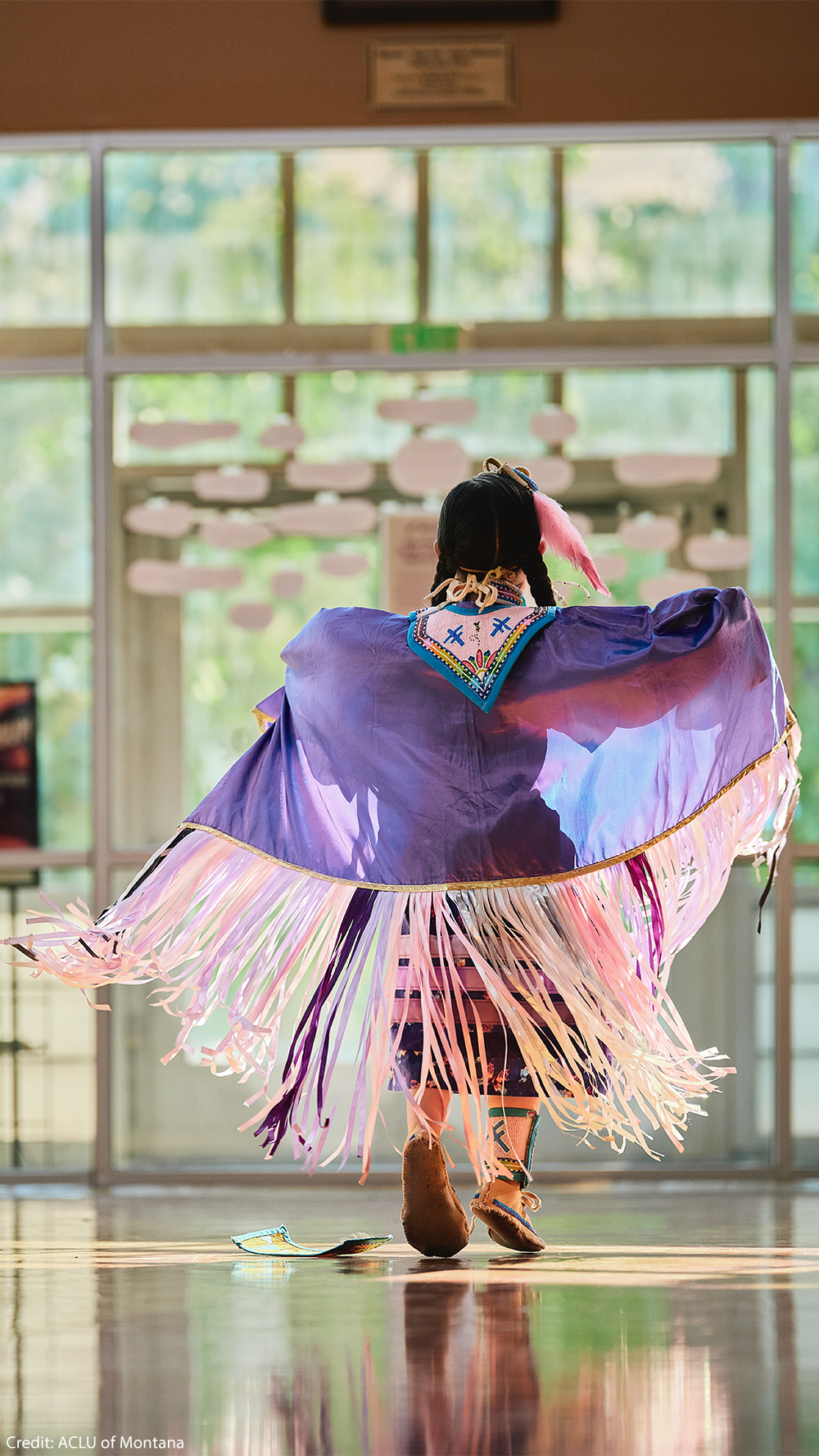 An Indigenous dancer is shown from behind performing indoors, wearing a flowing purple shawl with long pink, white, and purple fringe, intricate beadwork at the neckline, a pink feather in braided hair, and beaded moccasins. Sunlight streams through large windows and reflects off the polished floor.