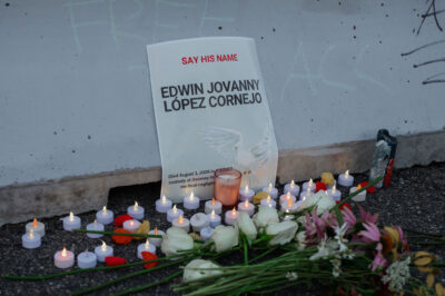 A memorial for Edwin Jovanny López Cornejo rests against a concrete wall, with a sign reading "Say His Name" above his name and noting that he died in ICE/GEO custody. White roses, other flowers, and dozens of small candles are arranged on the ground in front of the sign.