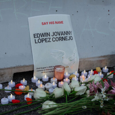 A memorial for Edwin Jovanny López Cornejo rests against a concrete wall, with a sign reading "Say His Name" above his name and noting that he died in ICE/GEO custody. White roses, other flowers, and dozens of small candles are arranged on the ground in front of the sign.