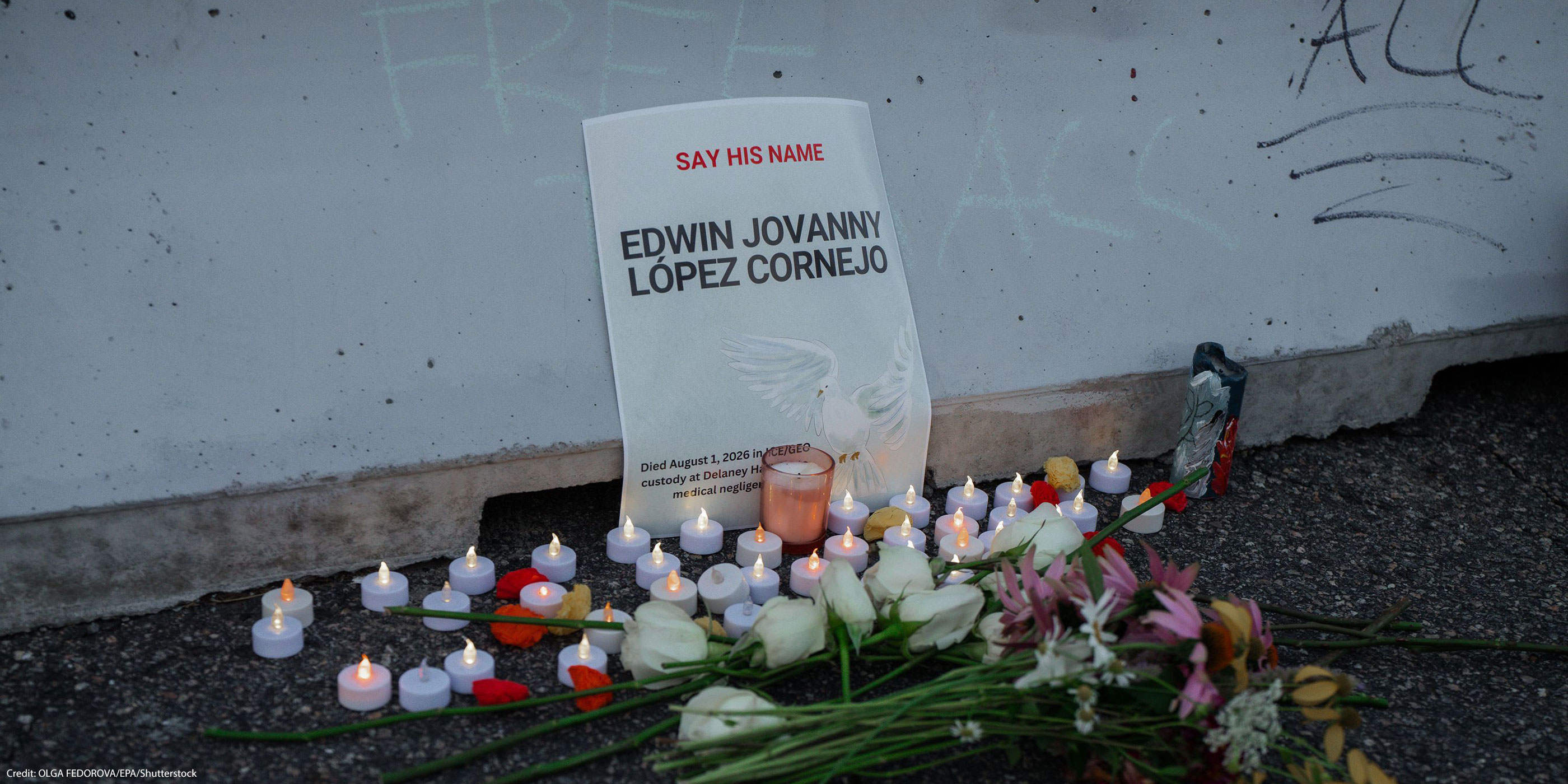 A memorial for Edwin Jovanny López Cornejo rests against a concrete wall, with a sign reading "Say His Name" above his name and noting that he died in ICE/GEO custody. White roses, other flowers, and dozens of small candles are arranged on the ground in front of the sign.