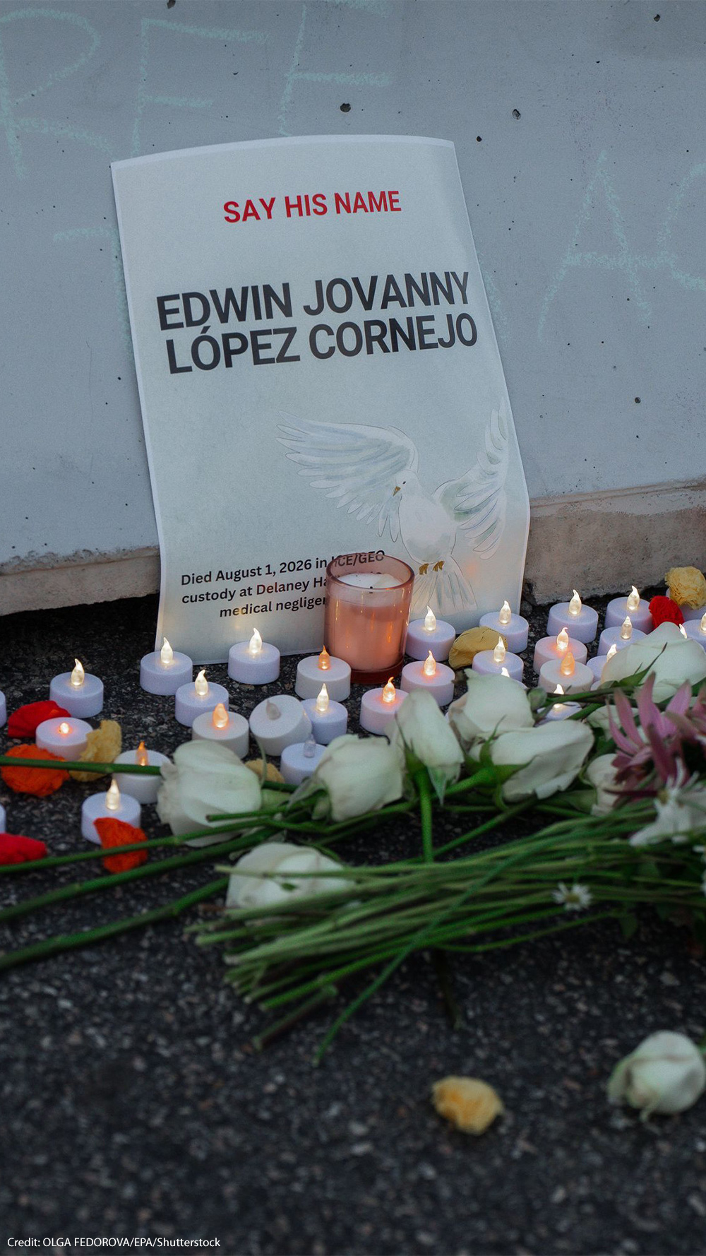 A memorial for Edwin Jovanny López Cornejo rests against a concrete wall, with a sign reading "Say His Name" above his name and noting that he died in ICE/GEO custody. White roses, other flowers, and dozens of small candles are arranged on the ground in front of the sign.